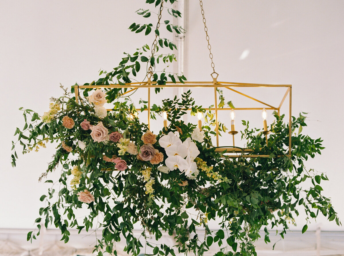 Hanging floral chandelier with greenery, roses, and white orchids suspended from a gold lantern-style frame inside a reception tent.