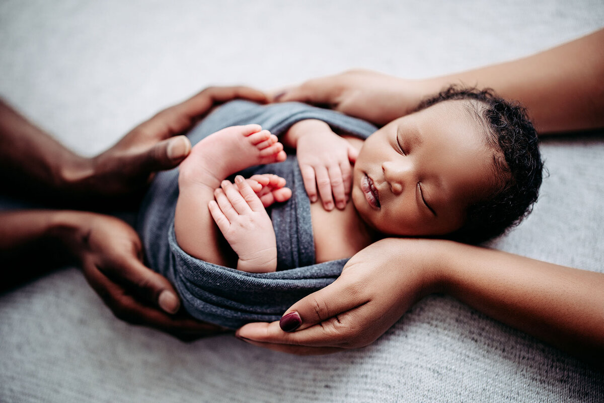 Swaddled newborn baby is held in mom and dad's hands during his newborn session at Jen Howell Photography in Germantown, TN near Memphis.