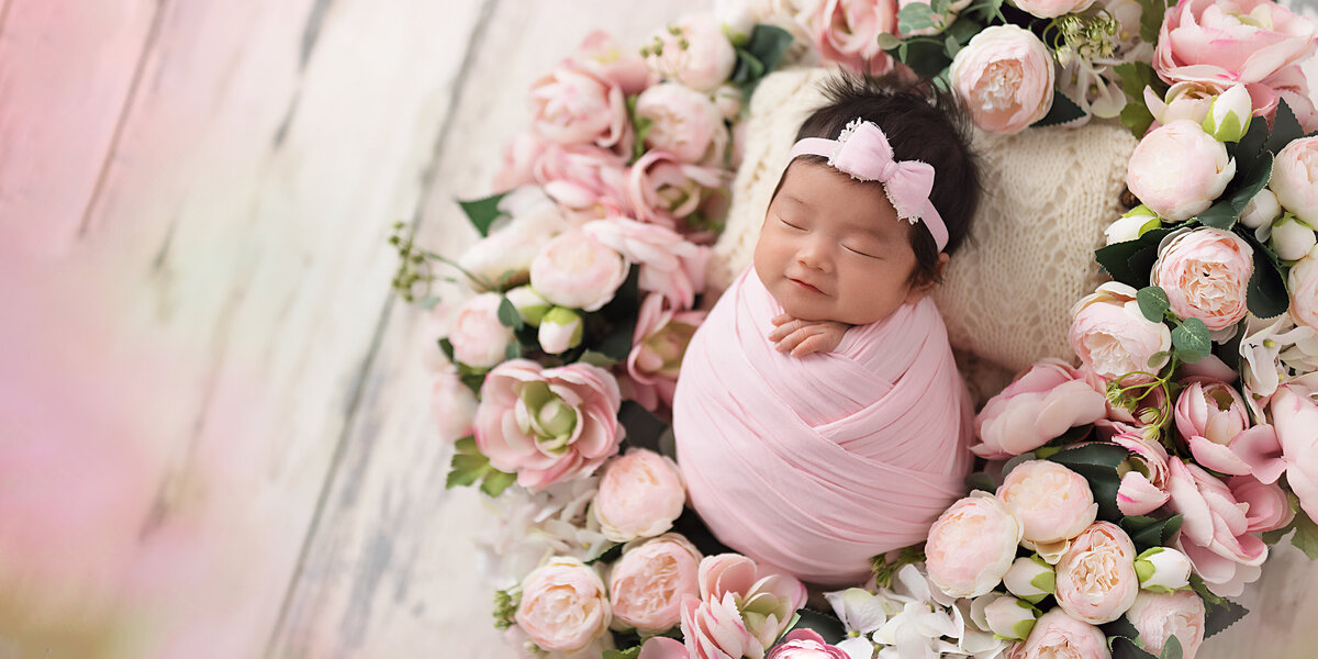 Newborn baby girl swaddled in pink and posed inside a circular wreath of soft pink peonies and florals on a light wood floor.