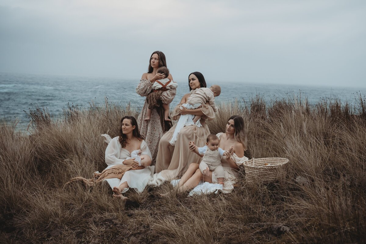 Five women dressed in neutral tones with their infants sit and stand in a grassy field beside the ocean, capturing tender moments during Wollongong motherhood sessions.