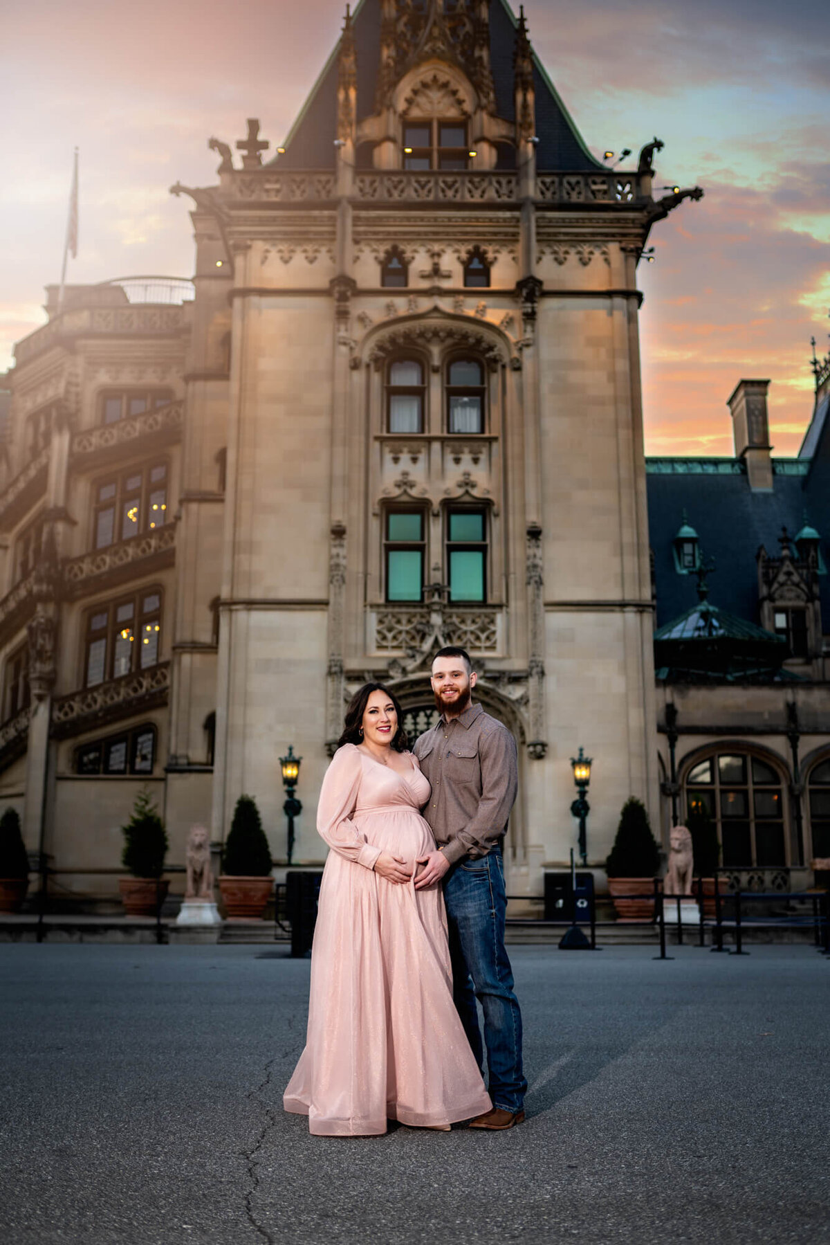 A beautiful mama to be in a long pink dress stands with her husband in front of the Biltmore Estate