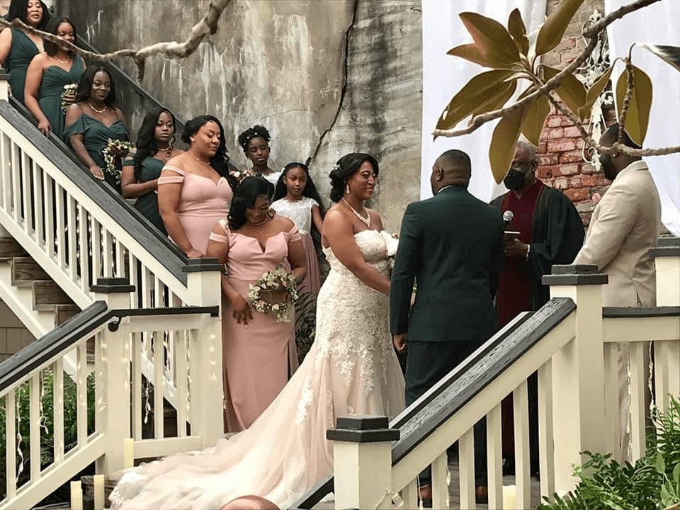 A bride and groom stand facing each other during their wedding ceremony on an outdoor staircase, surrounded by bridesmaids in pink dresses and groomsmen, with an officiant and lush greenery nearby.