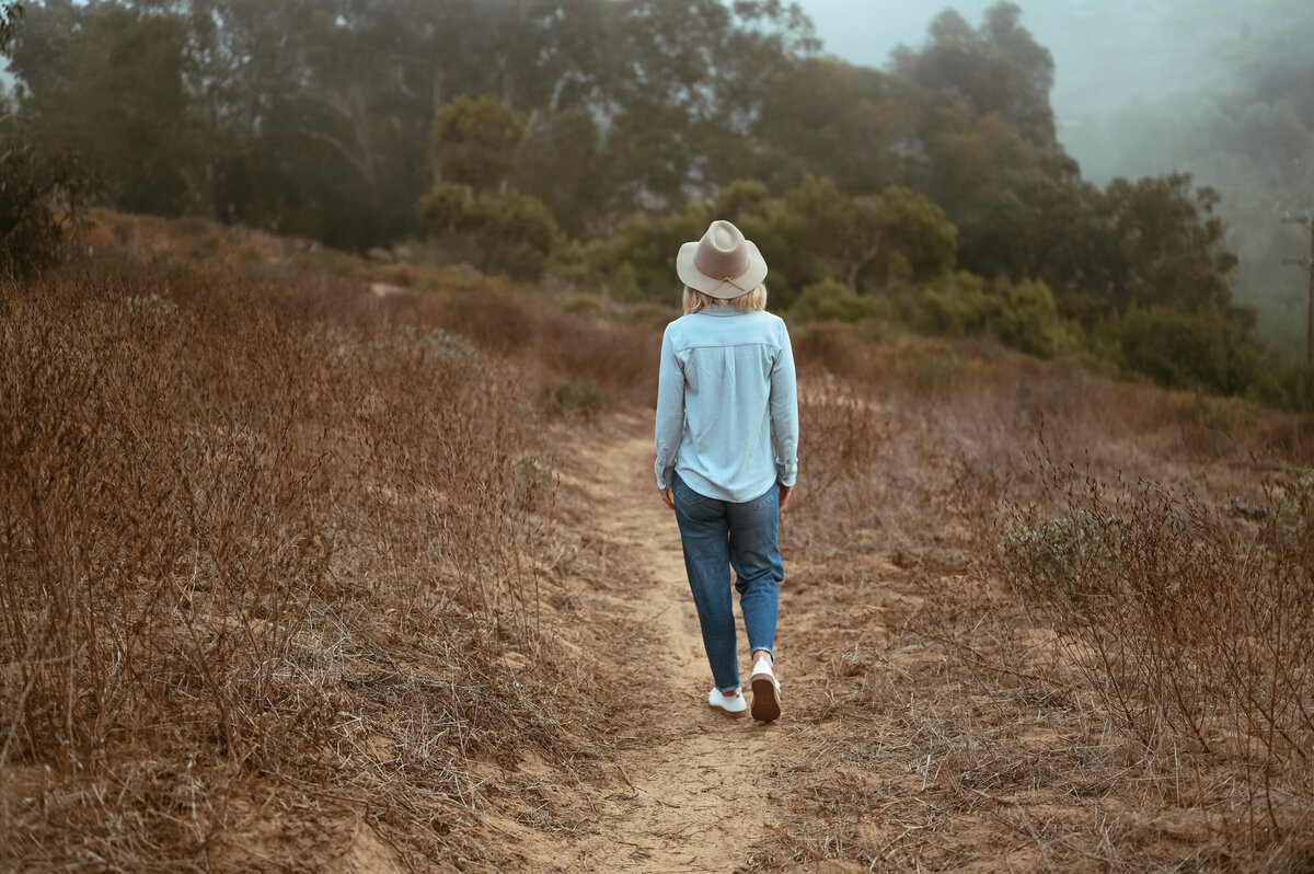 Woman walking alone on a nature trail in a hat and denim, representing calm and clarity.