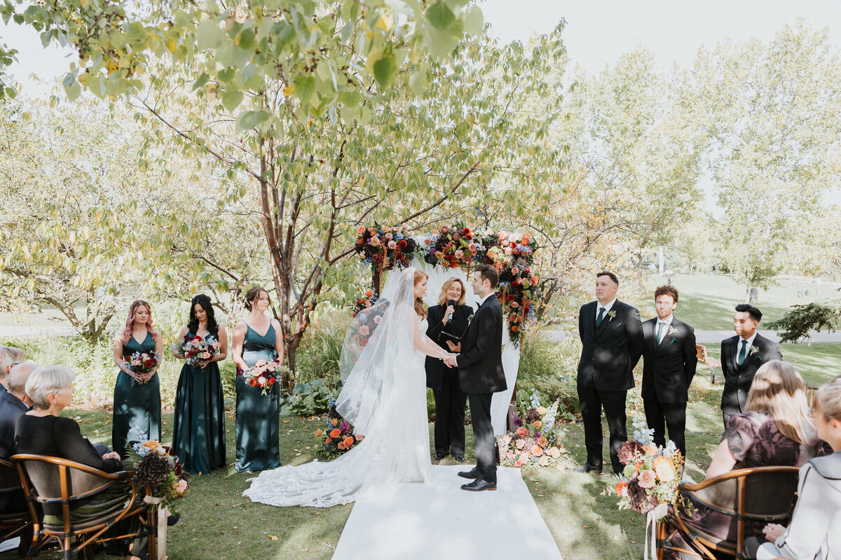 Outdoor wedding ceremony at River Café in Calgary, Alberta, featuring a couple exchanging vows under a vibrant floral arch. Surrounded by lush greenery, the bride in a flowing white gown and the groom in a classic black suit stand before their officiant. Bridesmaids in deep green dresses and groomsmen in black suits add to the elegant scene, while guests watch from floral-adorned wooden chairs. A romantic and picturesque setting along the Bow River.