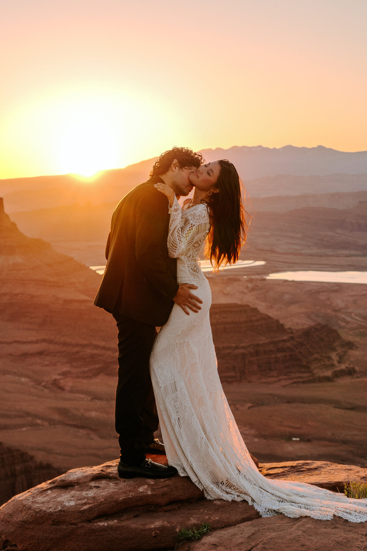 Bride and groom at Moab desert during sunrise the groom kissing her neck and embracing her while the sun rises in the background