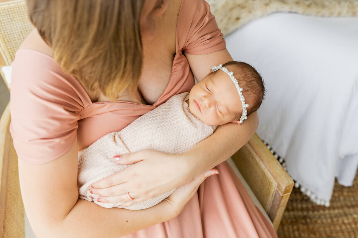 Mom in peach dress sitting in a chair and holding her newborn baby girl in a pale pink wrap and pearl headband