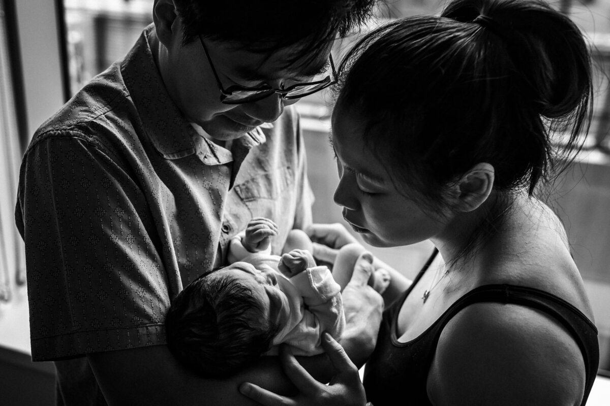 parents standing by hospital window holding newborn and looking down at him