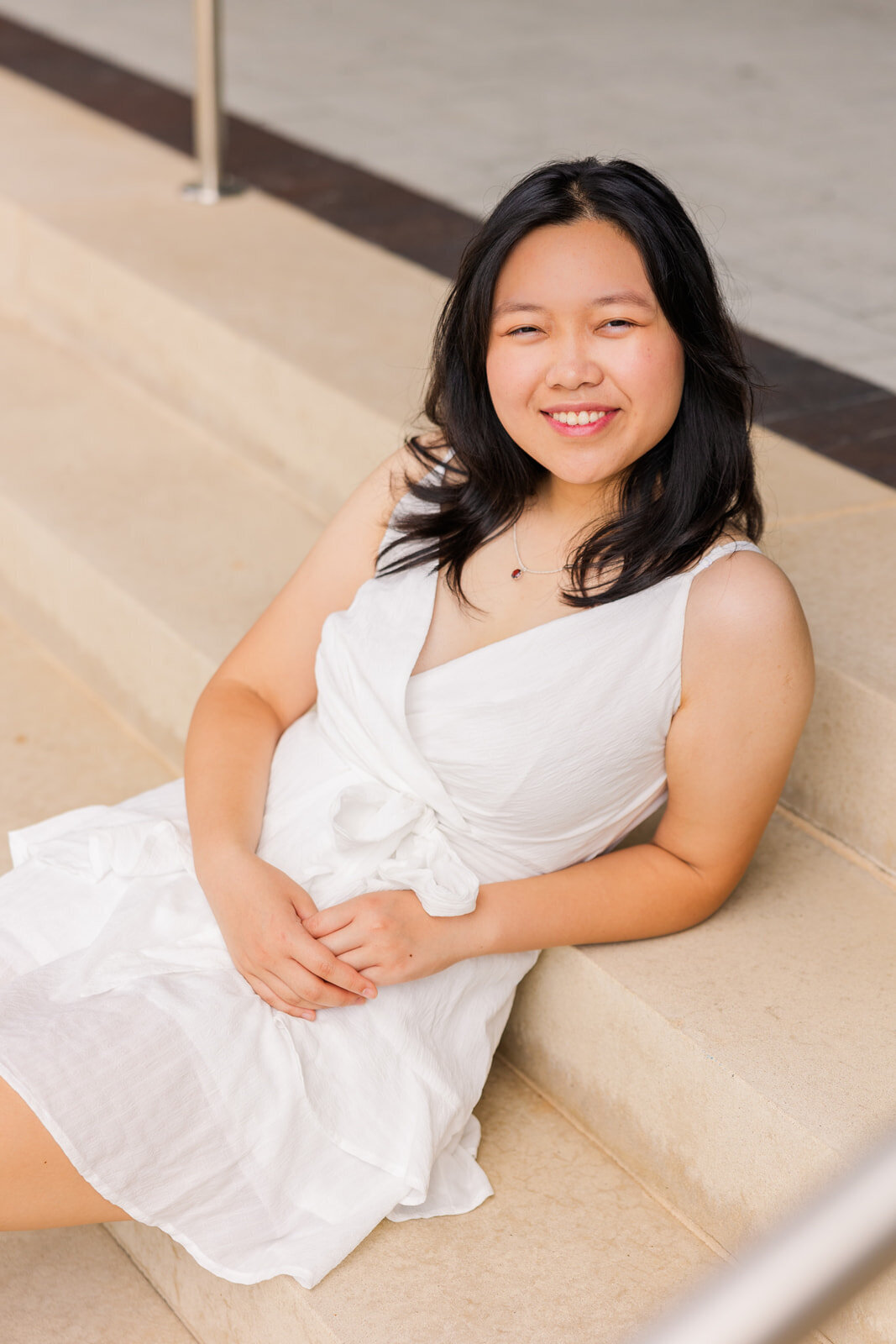 senior girl in white short dress sitting on stairs leaning back on her elbow during photo session with a neutral background in Sandy Spring