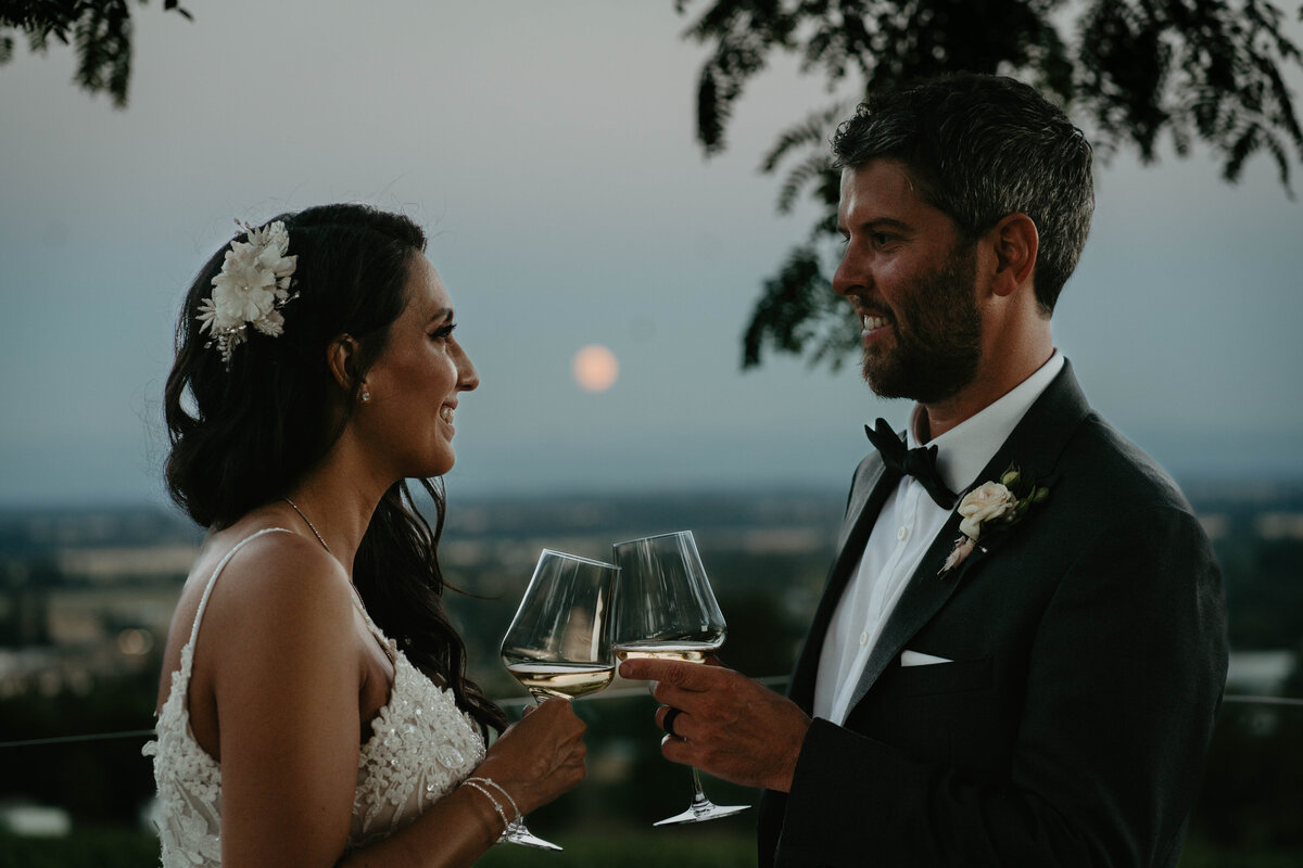 Bride and groom toasting at night with the moon in the background after their vineyard wedding