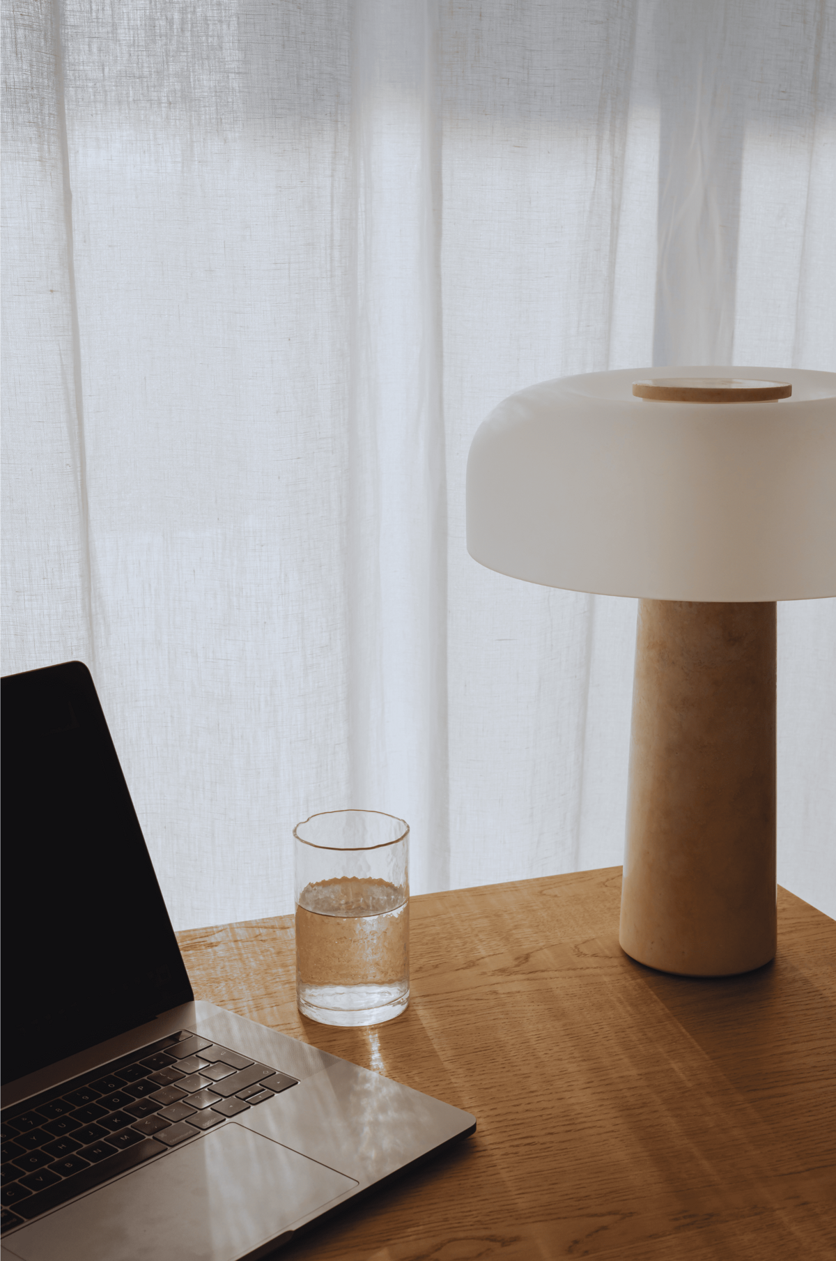 Egy minimalista home office beállítást mutat a képen: egy laptop, egy pohár víz és egy stílusos asztali lámpa egy fa asztalon. A háttérben függönyön átszűrődő természetes fény látható.