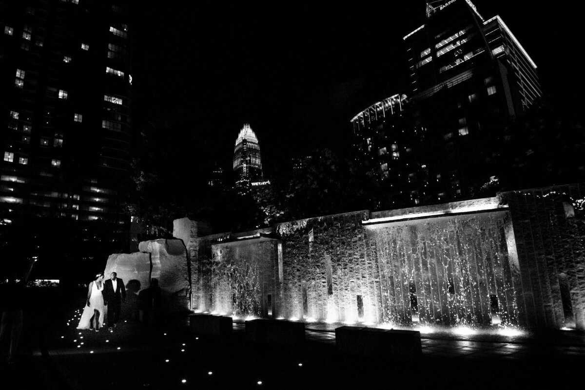 Black and white image of a black couple that just eloped walking through Romare Bearden Park at the waterfall with the Charlotte city skyline in the background by Charlotte wedding photographers DeLong Photography