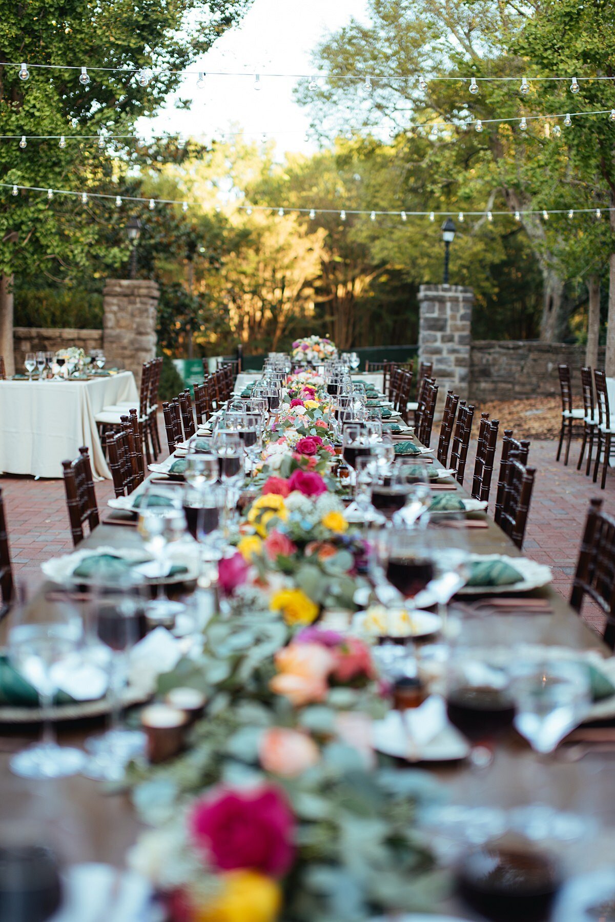 Very long table set for guests in the exposed brick courtyard at Cheekwood Botanical Garden. The dark wood farm table is set with hot pink and yellow roses in a thick green garland down the center of the table. The long reception table is set with gold chargers, forest green brocade napkins, glasses of water and glasses of red wine as well as matte gold flatware.