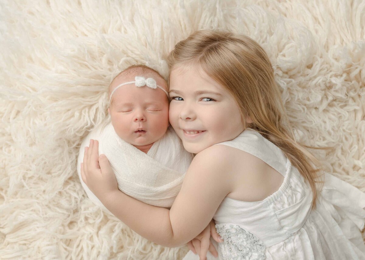 Big sister snuggling newborn baby laying on white flokati