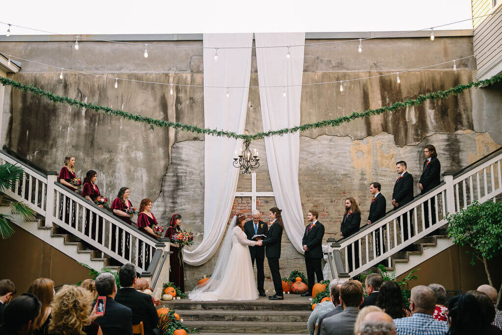 A couple stands at an altar exchanging vows, flanked by bridesmaids in burgundy on the left and groomsmen in black on the right, with guests seated below. The ceremony is outdoors, beneath draped white fabric and string lights.