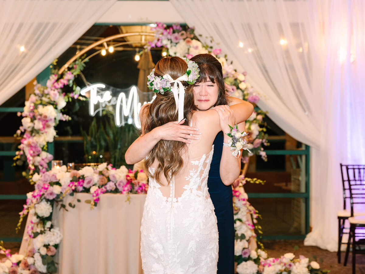 A bride in a lace dress and flower crown hugs her mother, in front of a floral arch. The setting is warmly lit and emotional.