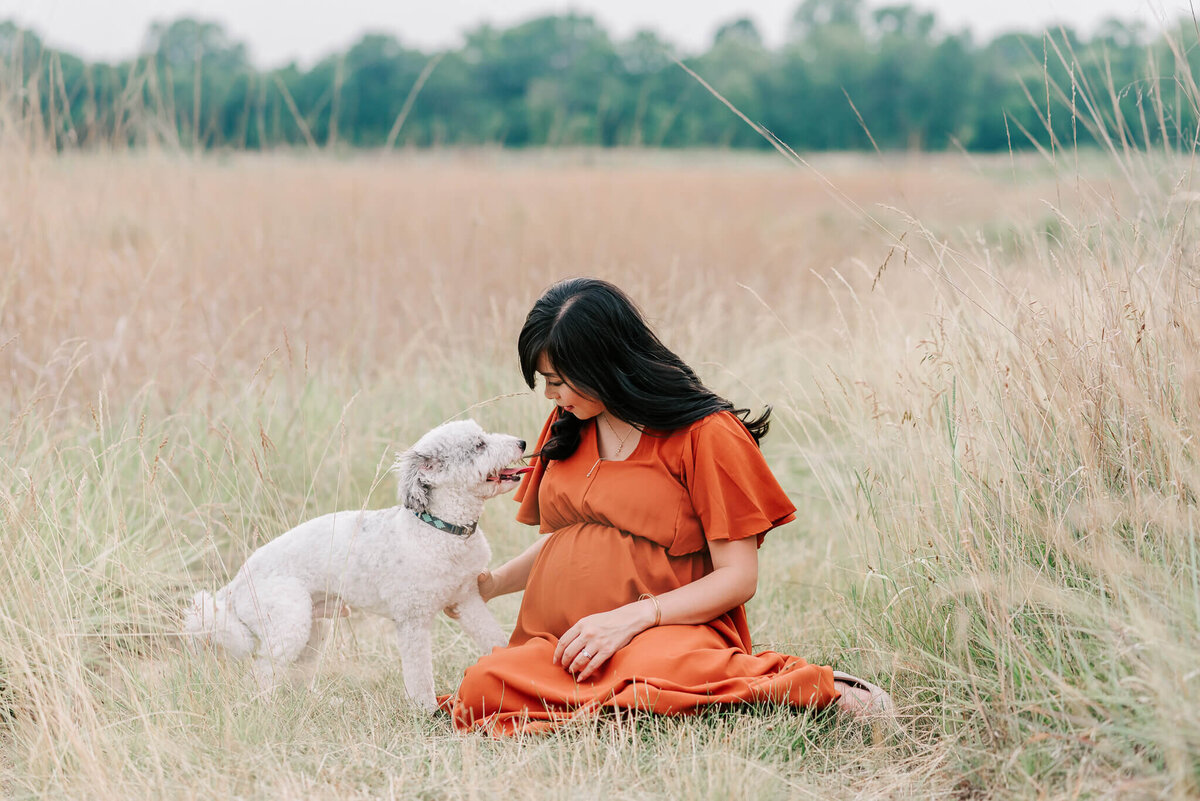 A pregnant woman sitting in the grass with her white dog during her maternity session
