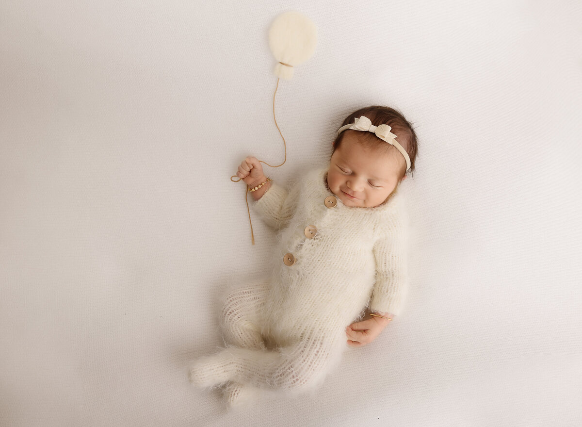 Baby girl is sleeping and smiling for her brooklyn newborn photoshoot. Aerial image. Baby is holding a felted balloon in one hand and is wearing a tiny bow headband. Captured by top Brooklyn newborn photographer Chaya Bornstein