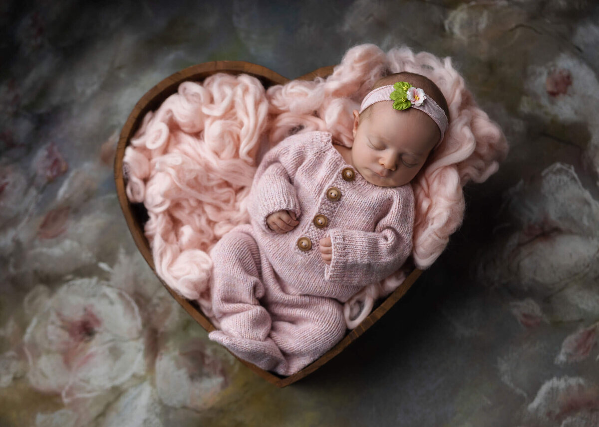 newborn baby wearing knitted pink romper and pink floral headband asleep in wooden heart shaped bowl