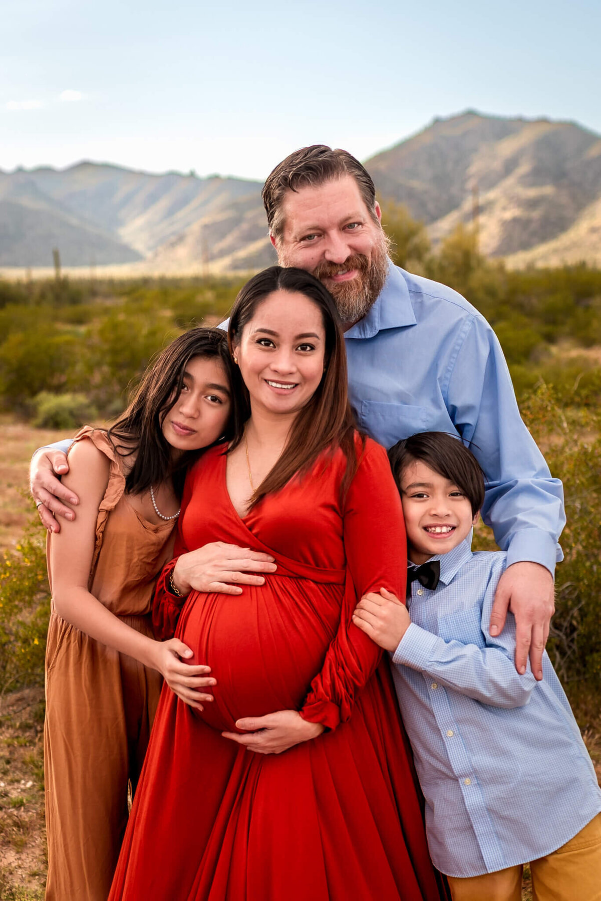 Glowing expecting mom smiling while family embraces her in Arizona desert. Portrait by Cactus & Pine Photography