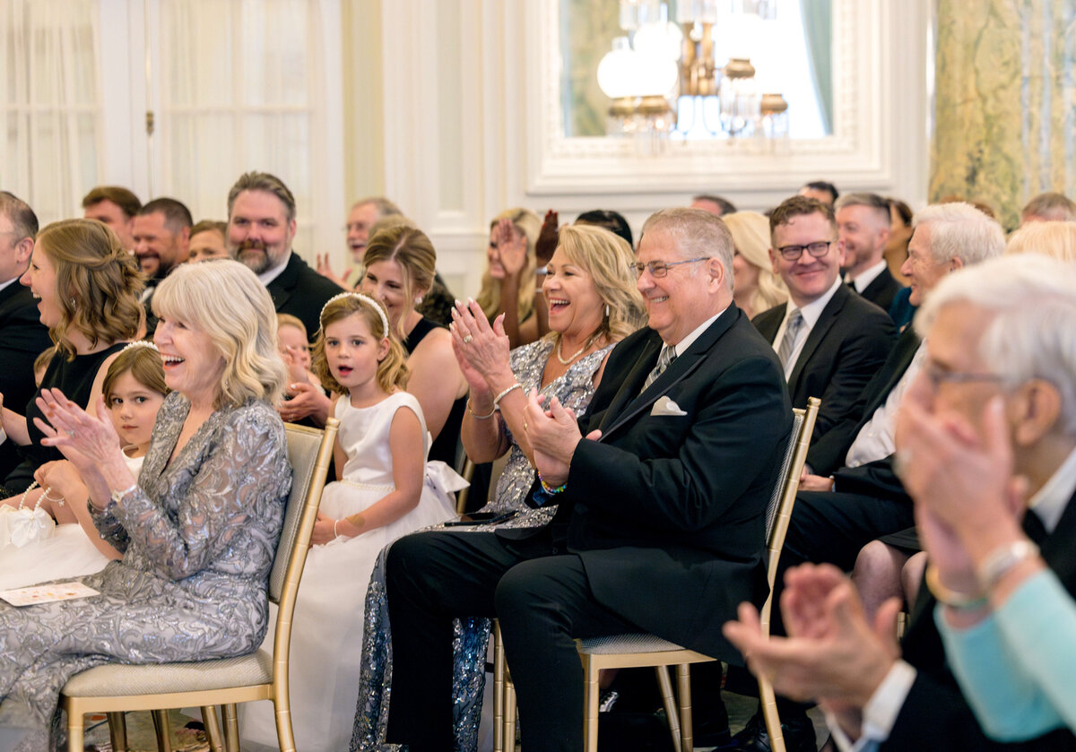 A smiling audience claps during an indoor event. They are dressed in formal attire, and include adults and children. The room is elegantly decorated with chandeliers and mirrors, creating a celebratory atmosphere.