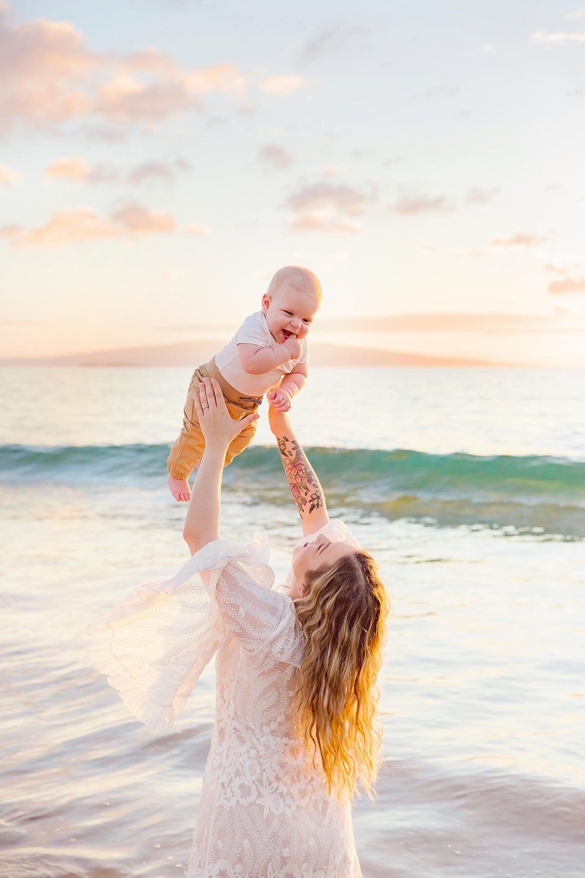 Young baby smiles as his mother lifts him over her head next to the ocean during their fun family photography session in Wailea