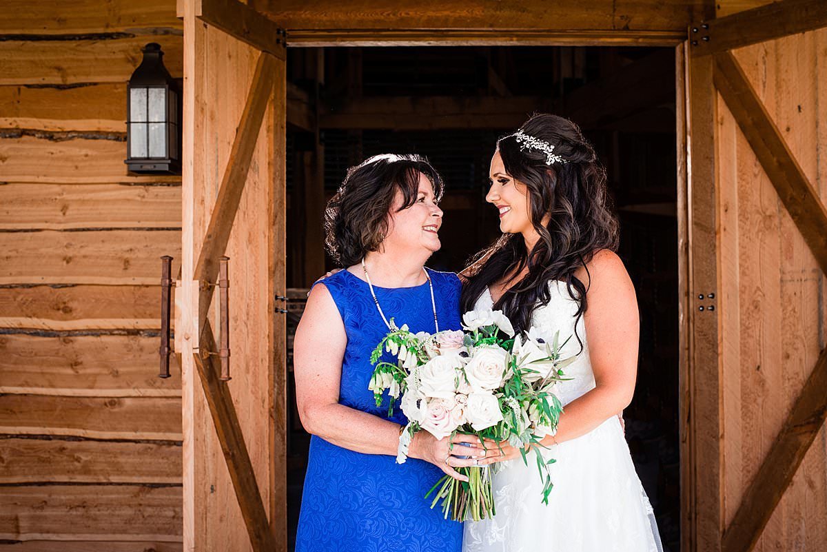 Bride and her mom smiling at one another for family portraits in front of barn wall