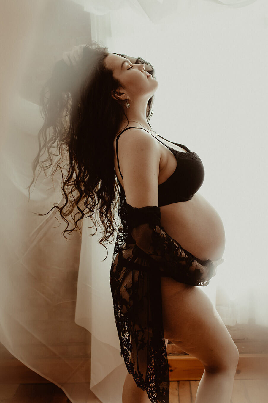 A beautiful pregnant woman wearing a black bra and panty set with a black robe standing in front of a window fixing her hair