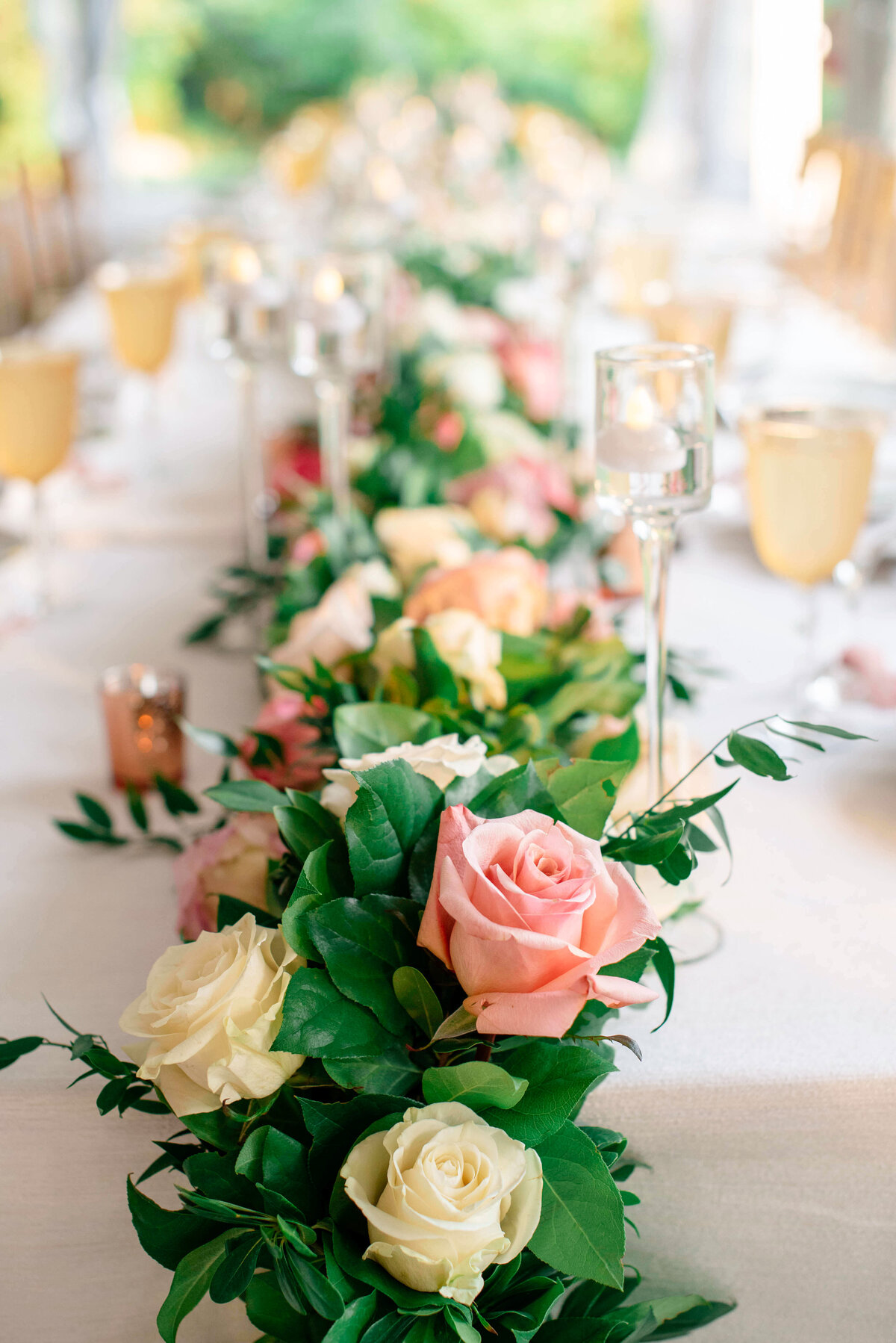 A long table decorated with an elegant floral arrangement of pink and white roses and green leaves. Tall candle holders are interspersed along the centerpiece. Golden-toned glasses are placed beside each setting on a white tablecloth.