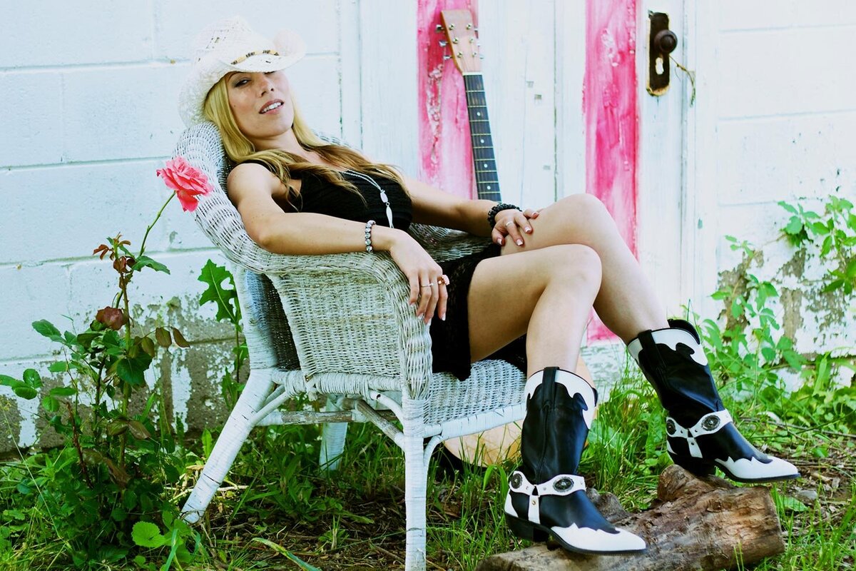 Female Country Music Artist Portrait Krysta Lee sitting leaned back in white wicker chair wearing black dress cowboy hat and black and white cowboy boots