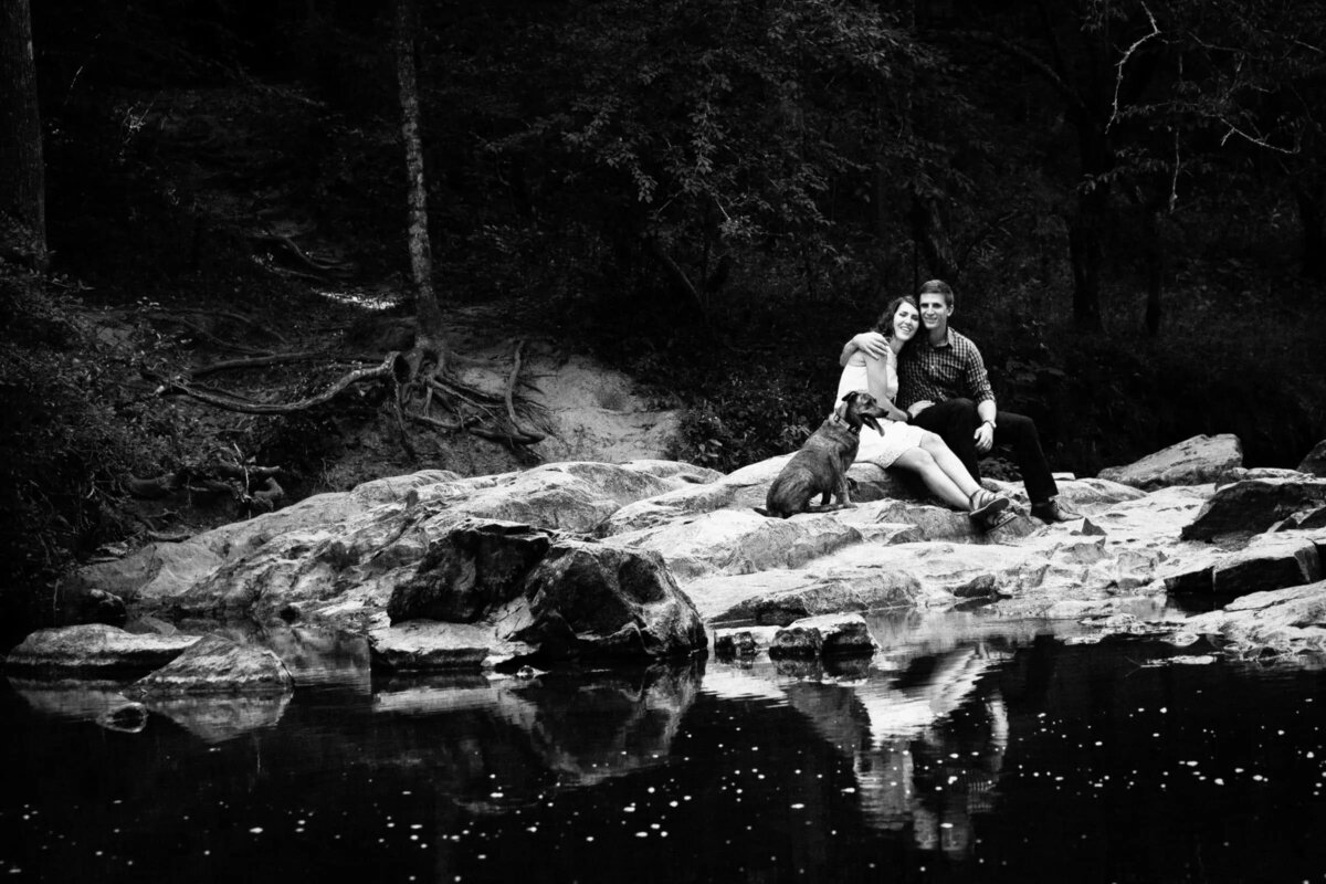 Monochrome photo of a couple sitting closely on a rock by a reflective water body, surrounded by a forest