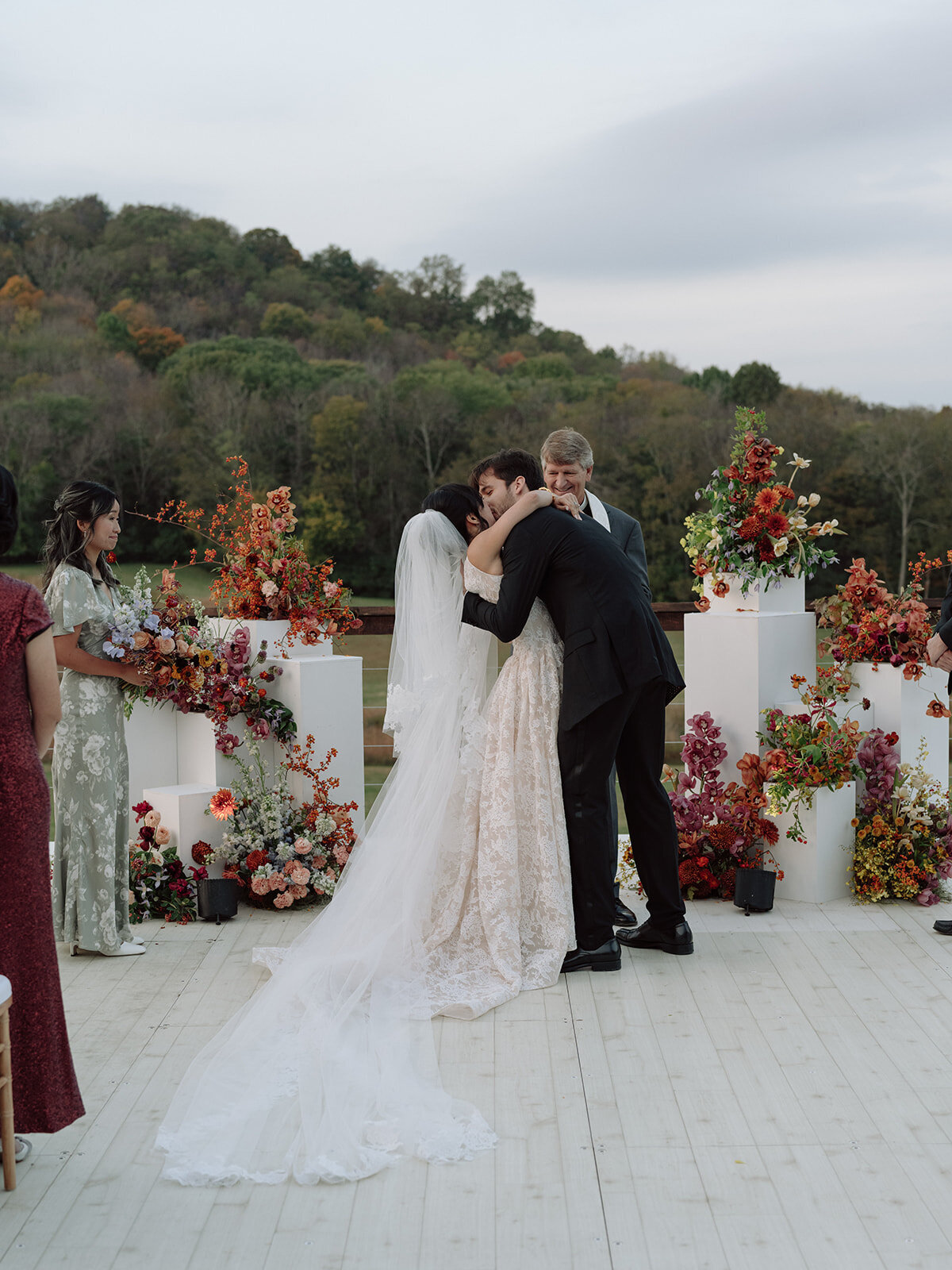 Sculptural columns wedding ceremony backdrop. Stair step white ceremony columns adorned with sculptural floral meadows in fall hues. Clear top tent build out for fall wedding reception in Tennessee hills. Wire threaded orchids, bittersweet berries, butterfly ranunculus, and tulips are highlighted in this wedding ceremony backdrop. Artistic fall color palette in copper, gold, ruby, lavender, and rust. Design by Rosemary & Finch Floral Design in Nashville, TN.