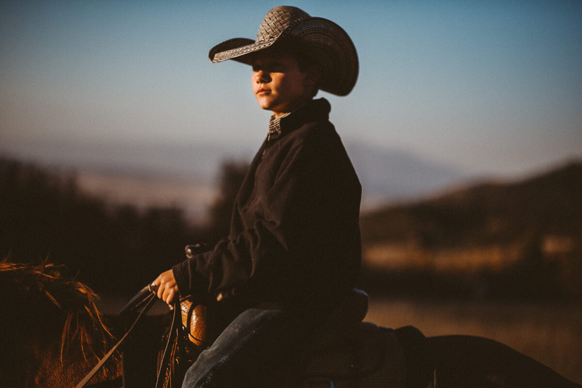 boy sits on top of horse on his family's ranch in colorado as the sun sets in the background sky