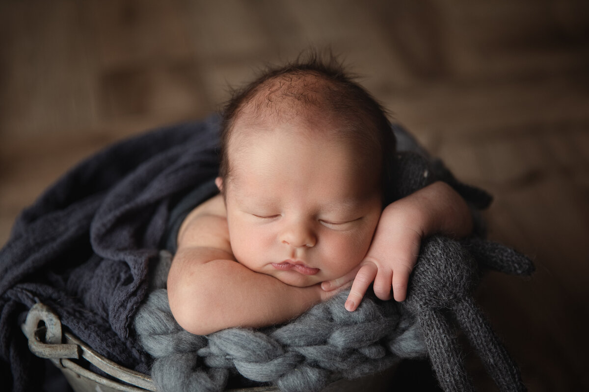newborn baby boy posed in a rustic tin bucjet holding a grey bunny  looking peaceful and asleep