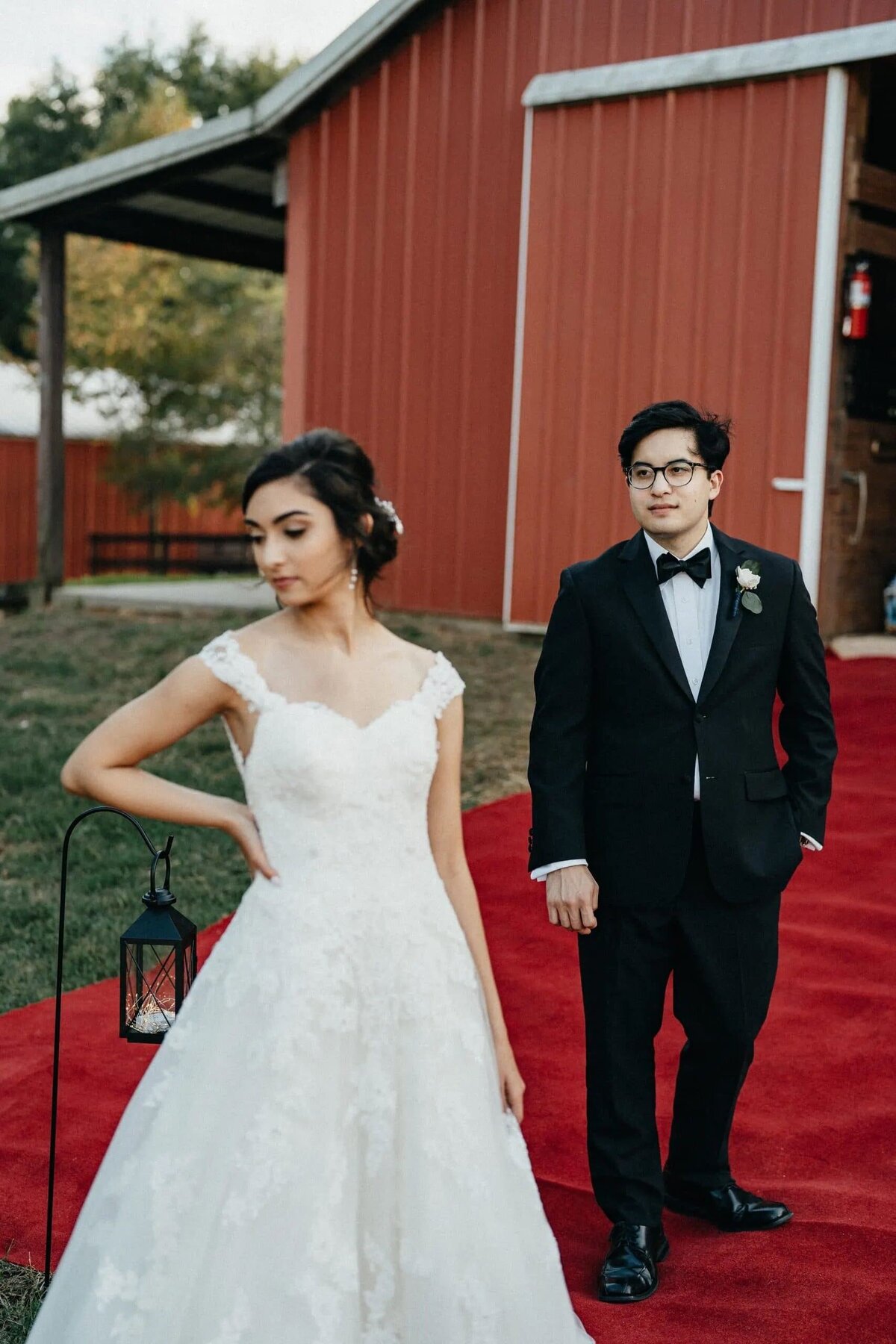 A bride and groom on a red carpet outside a barn venue, the bride looking down thoughtfully, and the groom looking towards the camera.