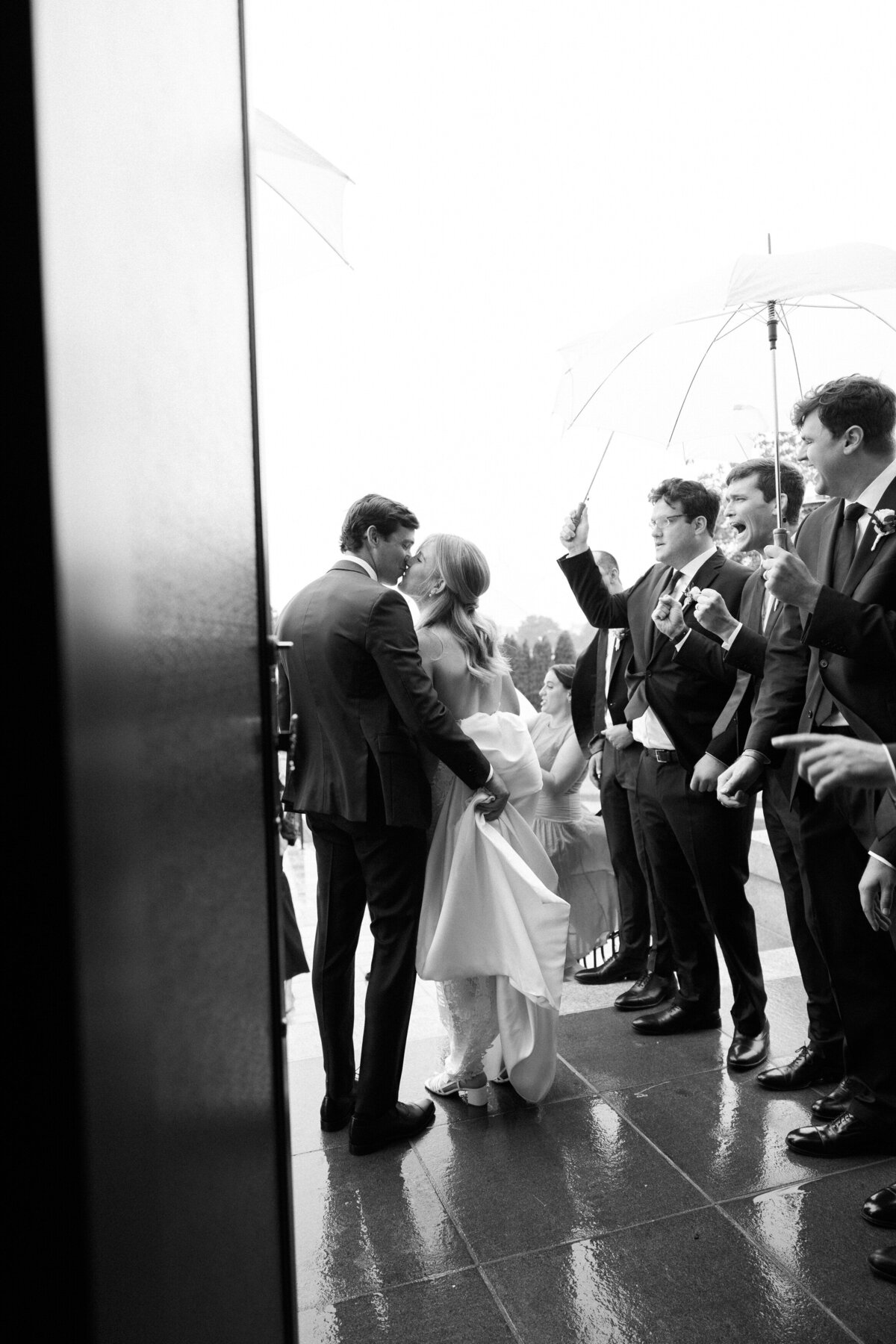A black and white image of the couple sharing a kiss as they leave the church, surrounded by cheering groomsmen holding umbrellas. The groom lifts the train of the bride’s dress as they step onto the wet pavement.