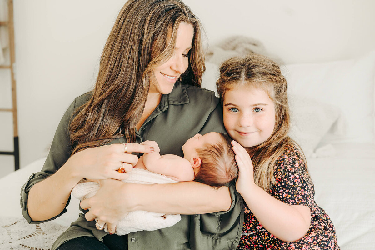 older sister smiles at camera while snuggling her baby sister for newborn photos