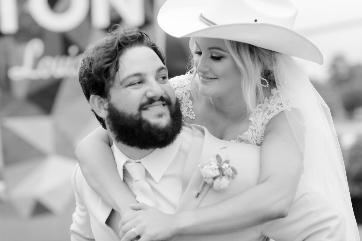 bride on her grooms back with her arms around his shoulders while looking down at him while wearing the grooms cowboy hat
