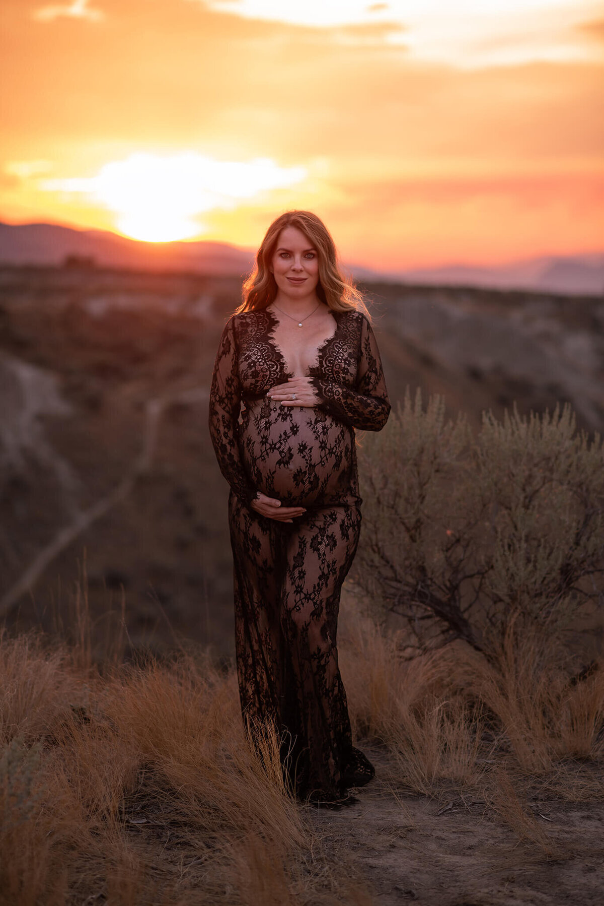 A woman wearing a sheer black lace dress stands against a dramatic sunset background while she holds her belly and smiles. Taken by Brittany Danielle Photography