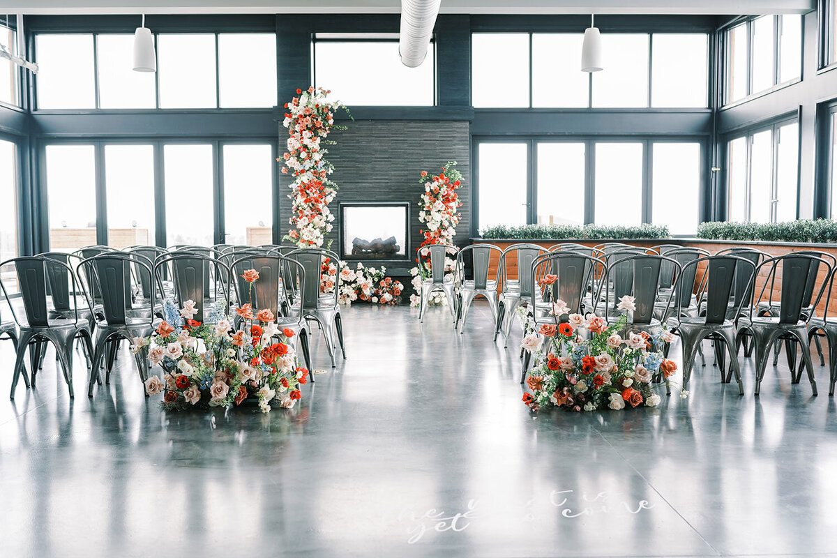Indoor wedding ceremony setup with modern metal chairs, colorful floral aisle arrangements, and a bold floral installation framing the fireplace.