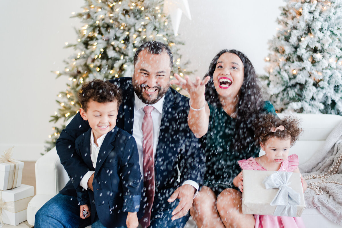 family of four sitting on a couch with presents in front of the christmas tree snow falling by miami christmas mini session photographer msp