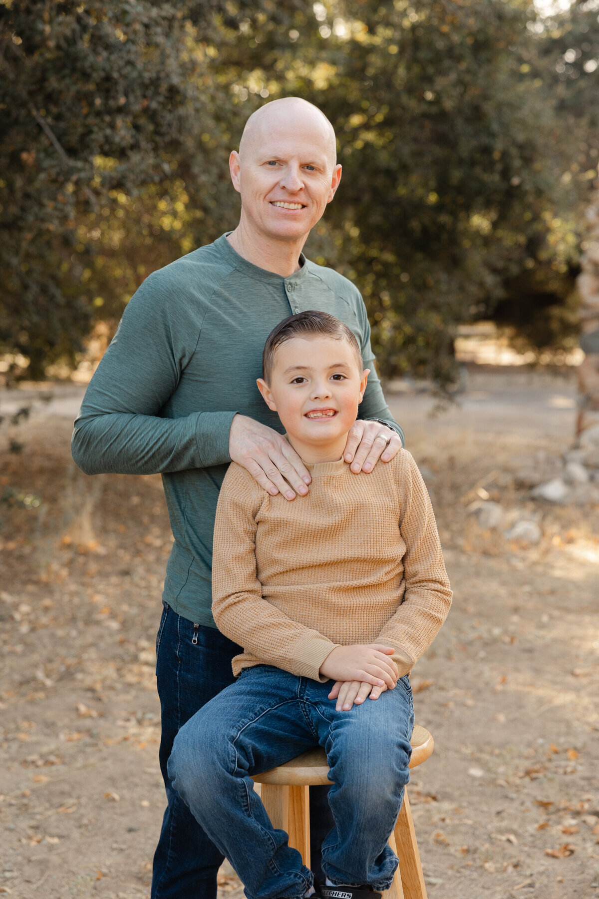Father standing behind his young son seated on a wooden stool outdoors, both smiling in earthy fall outfits. Photographed by Yucaipa family photographer Kaitlyn Dawn Photography