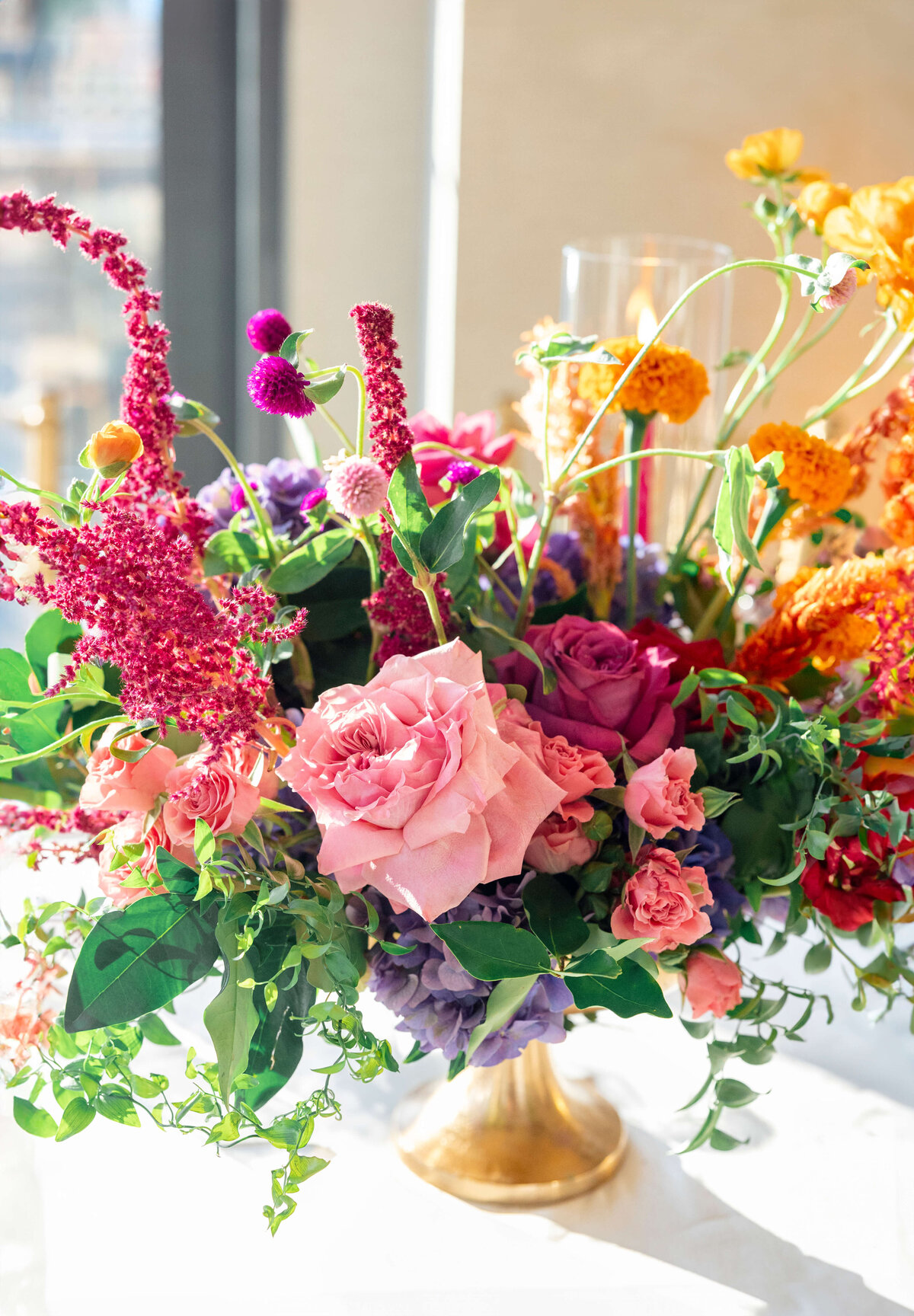 A vibrant floral arrangement in a golden vase, featuring pink roses, purple blossoms, and orange and yellow flowers with green leaves. A lit candle in a glass holder is nestled among the flowers.