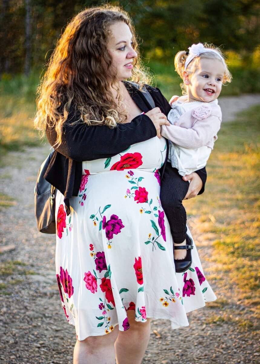 a pregnant woman in a white floral dress holding her toddler daughter