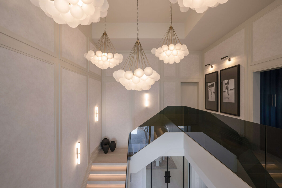 The top of a modern white staircase, bordered by a sleek, black glass railing on the right side. The staircase leads to a large, open landing, with the black glass railing continued through the hallway. Above, bold, bubbly chandeliers emit a soft light and add a contemporary style to the space.