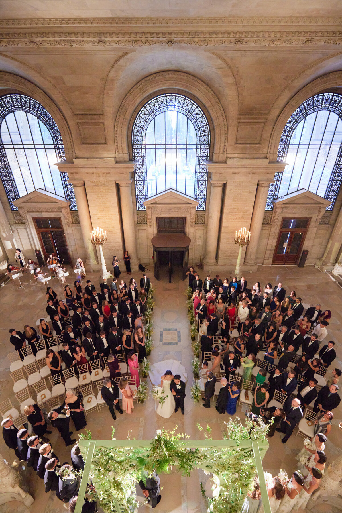 This is a stunning photo captured at the beautiful New York Public Library in New York City by talented photographer Shawn Connell, with videographers Aaron Navak Films. It showcases a wedding ceremony inside a grand hall with large arched windows and chandeliers. Guests are elegantly seated on both sides of a central aisle, creating a graceful semicircle around the couple standing under a romantic floral archway. The venue's elegant and classical architectural style adds a touch of timeless sophistication to the occasion.