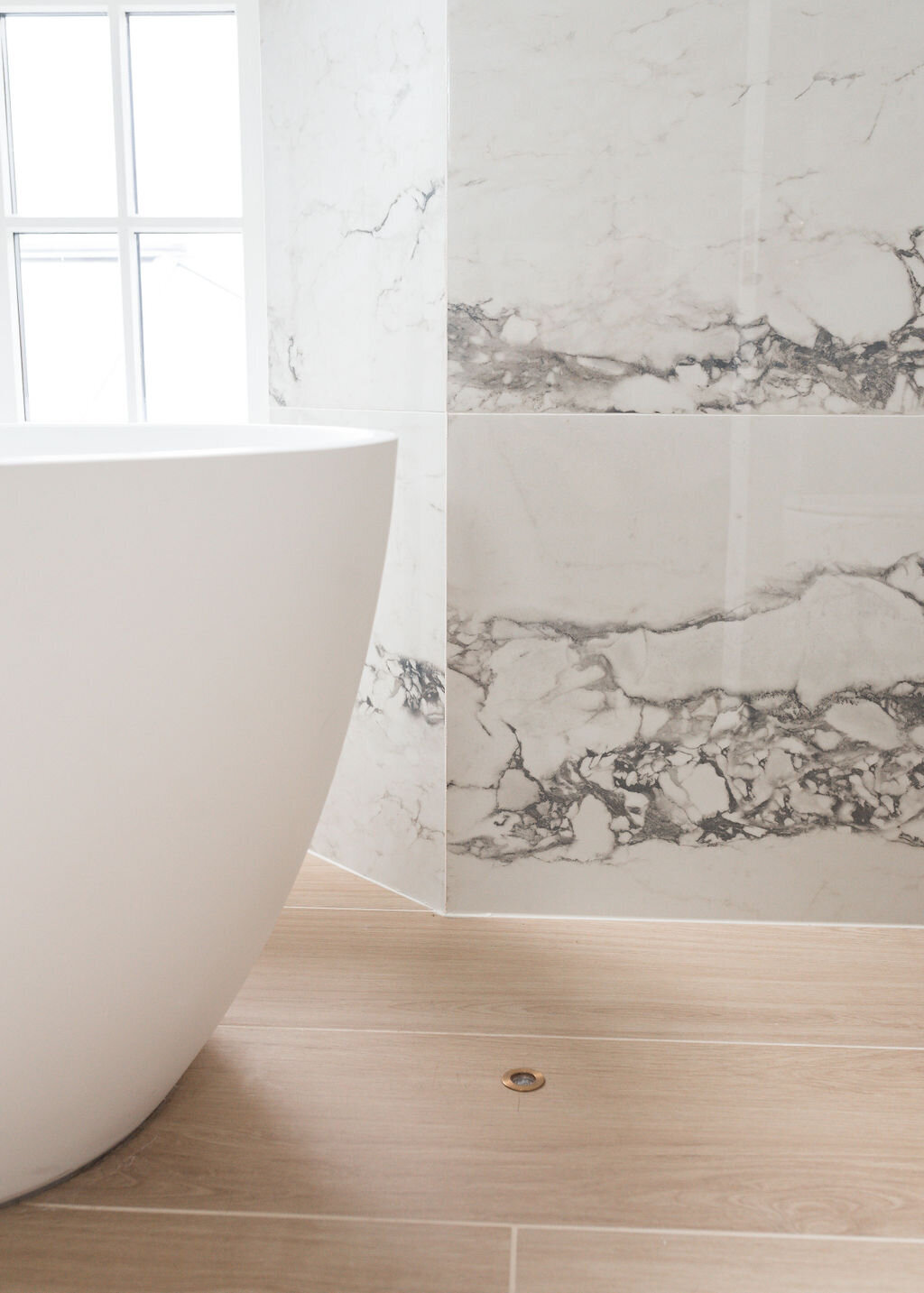 Close-up view of the corner of a modern white bathtub in a modern, grey marbled bathroom with light wooden floors.