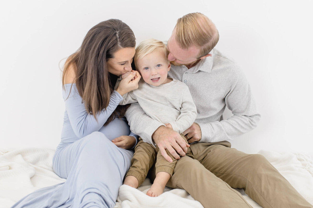 A family of three sitting on a soft blanket in a bright setting. The woman, in a light blue dress, and the man, in a gray shirt, lovingly embrace a smiling young child between them. The mood is warm and joyful.