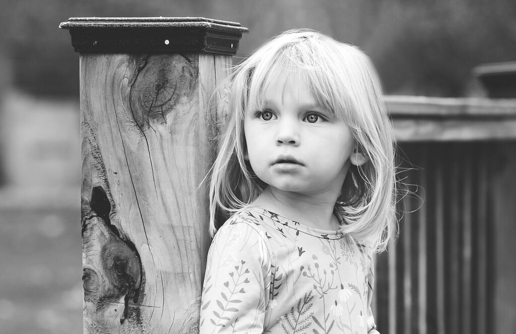 Toddler girl leans against fence post looking off into the distance.