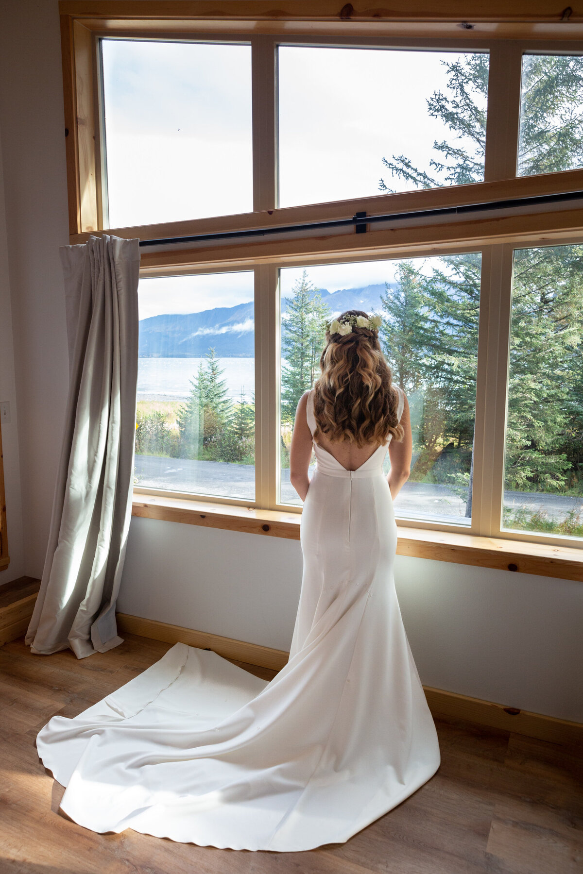 A bride stands looking out the window at her airbnb in Alaska.