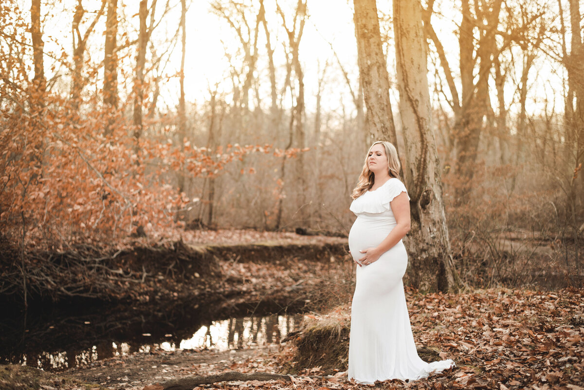 Pregnant woman at 34 weeks in outdoor fall setting wearing white maternity gown and holding baby bump